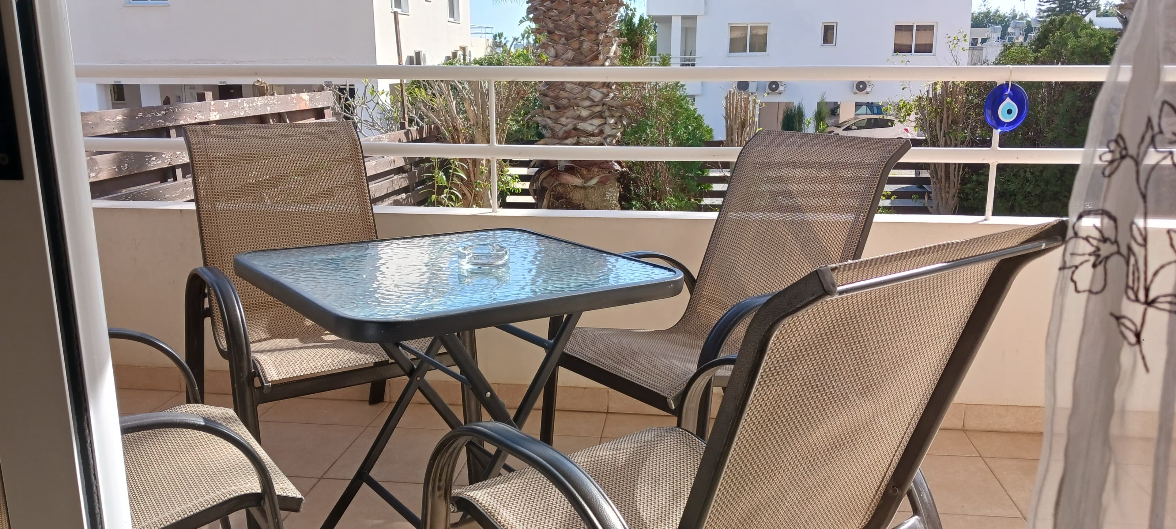 Sunlit covered terrace with a bistro table and chairs beside a palm tree at the Nissi 3 garden apartment