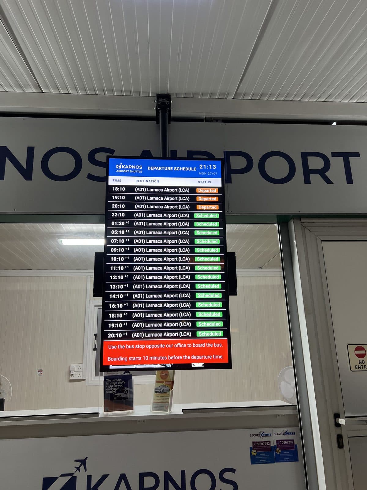 Physical Kapnos Airport Shuttle departure board in Ayia Napa listing services to Larnaca Airport, photographed 27 July 2026